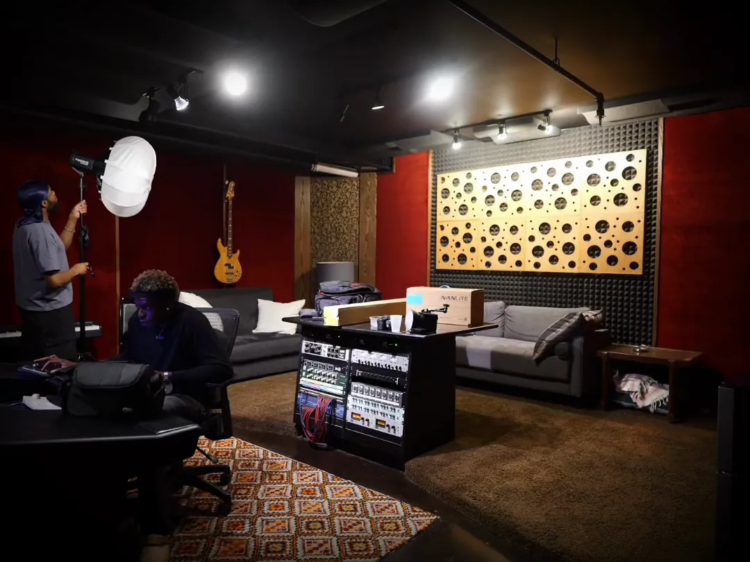 A red-walled recording studio: mixing console, couches and a lit acoustic panel, the lights dropping to one lamp.