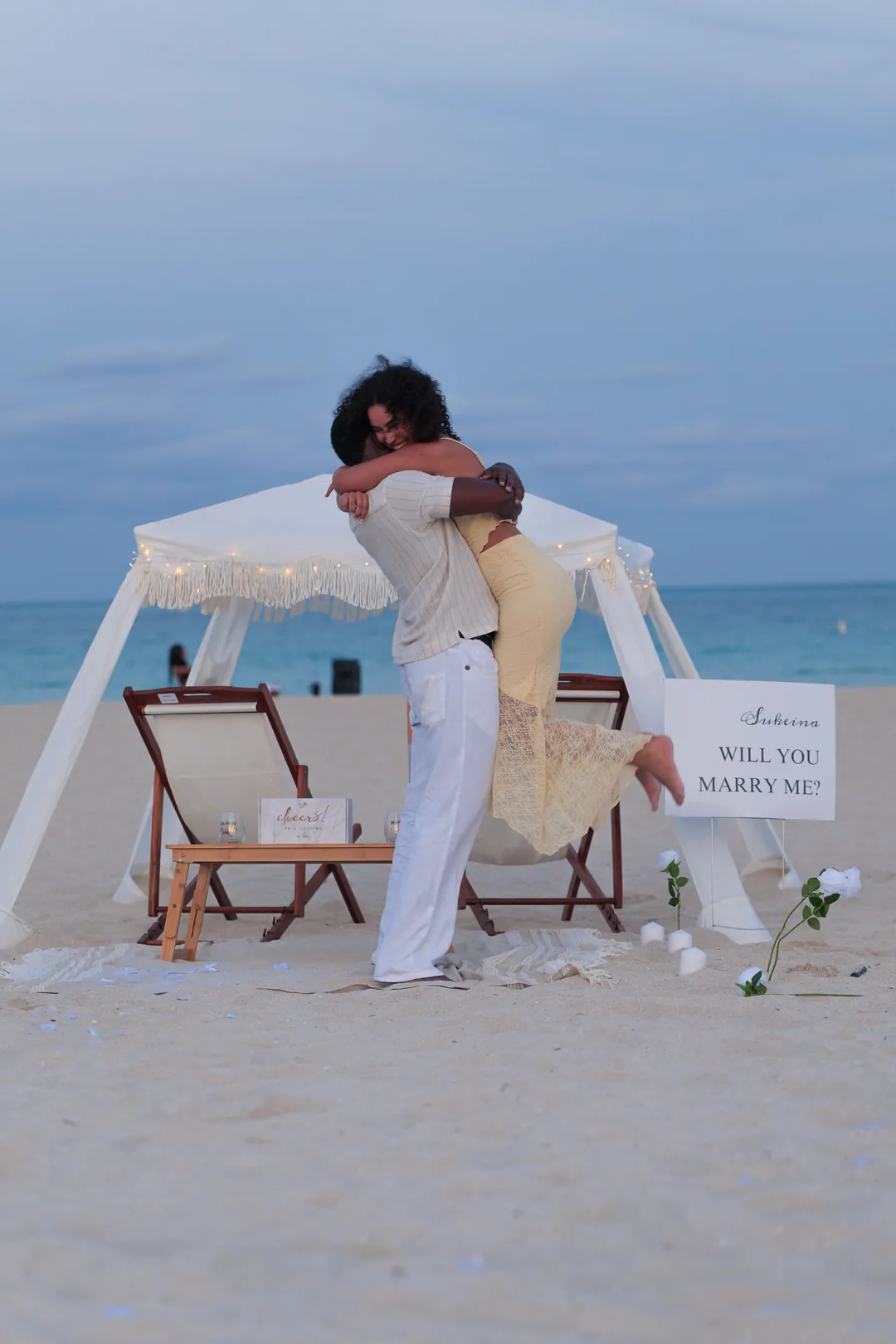A woman leaps into her partner’s arms on the beach after a proposal, a “Will you marry me?” sign beside them.
